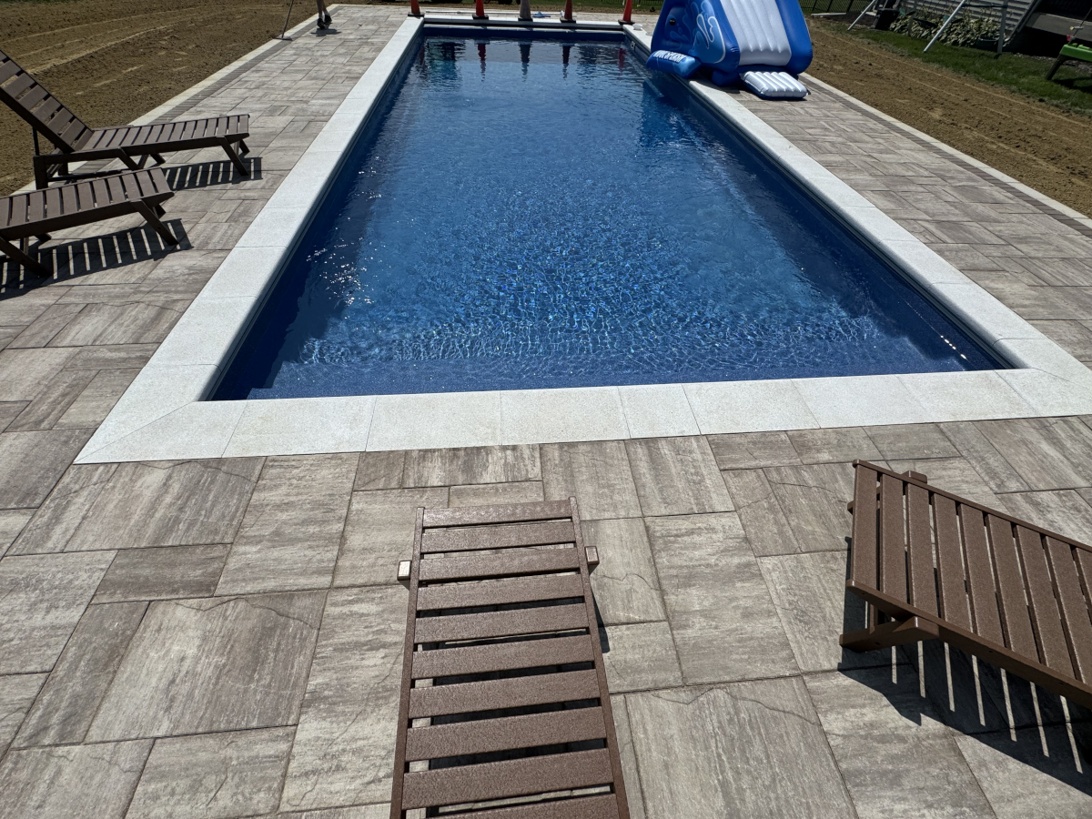 Rectangular pool with blue water, surrounded by beige stone tiles. Wooden loungers are placed along the sides, and a blue inflatable slide is at the far end.